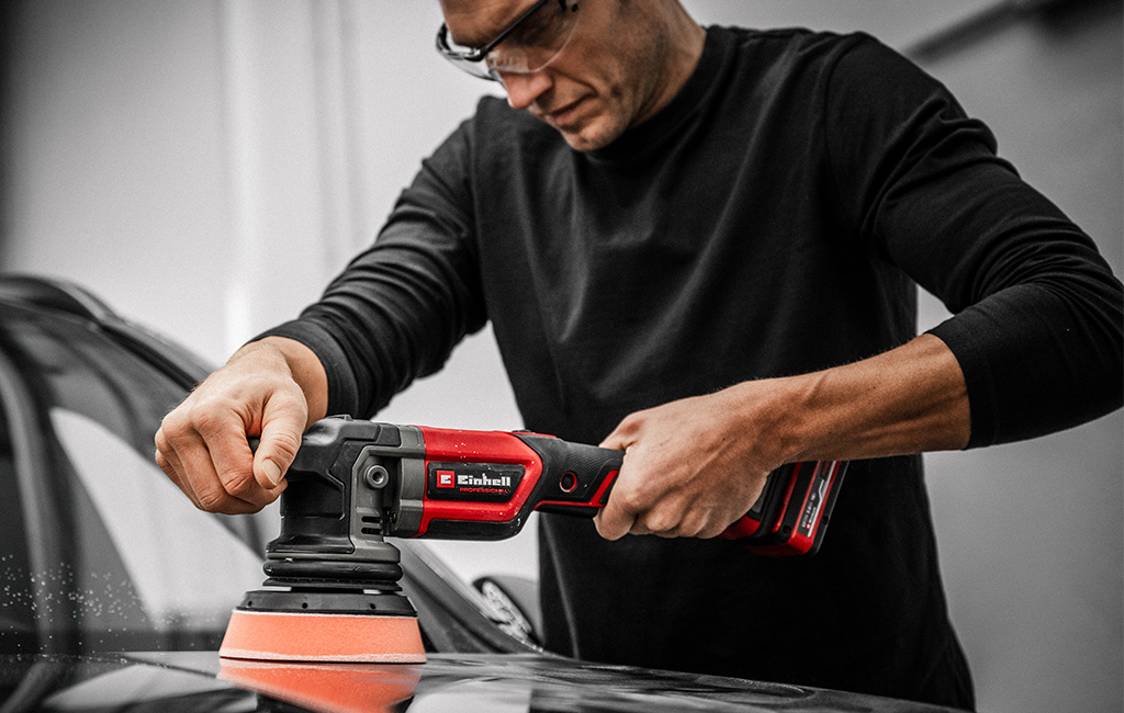 A man positions an Einhell cordless polisher on car paint, close-up in a workshop.