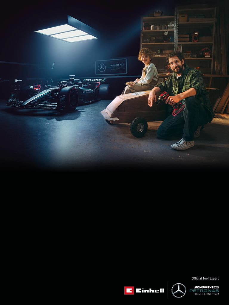 A father and child in a workshop with a homemade soapbox car. In the background, a Mercedes-AMG PETRONAS Formula One car and Einhell tools.