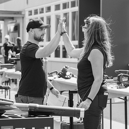 Un barbat si o femeie fac un High-Five la un eveniment Einhell in aer liber cu exponate de unelte, alb-negru.