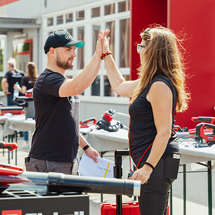 Un barbat si o femeie fac un High-Five la un eveniment Einhell in aer liber cu exponate de unelte, fotografie color.