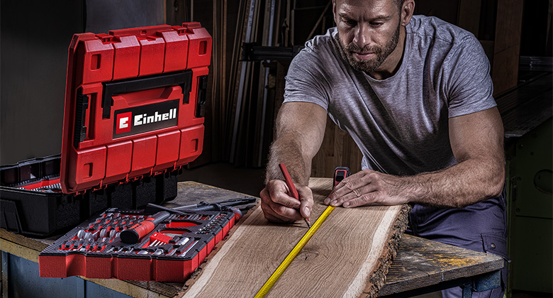 A man measures a wooden board with a tape measure next to a Einhell tool case set.