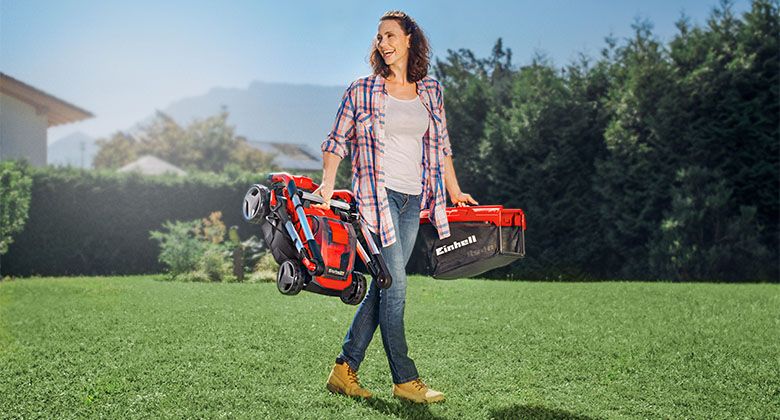 Woman carrying her folded cordless lawnmower and grass catcher through the garden.