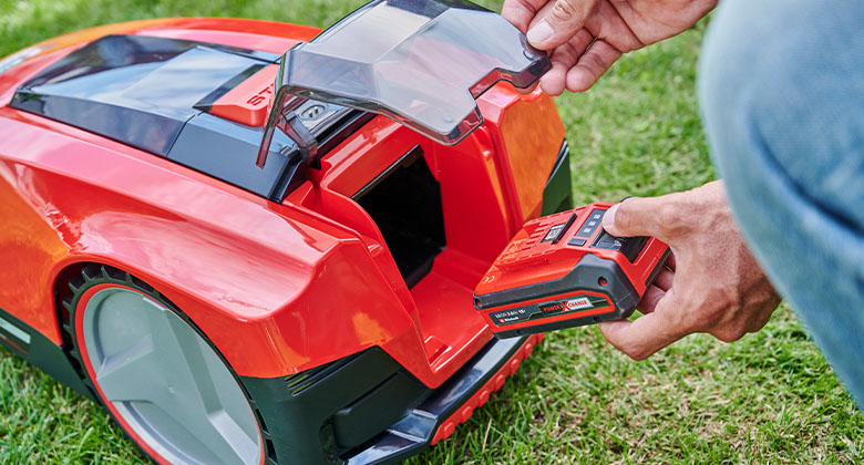 A person is inserting a red rechargeable battery into a red robotic lawnmower using a transparent battery compartment lid.