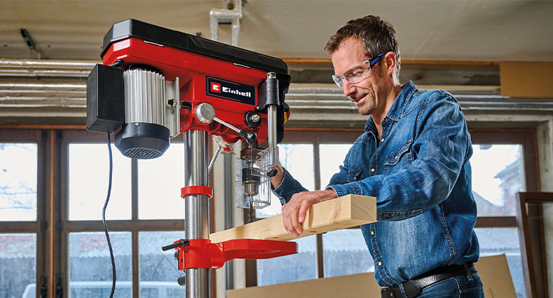 A man using a bench drill to drill a hole in a thick wooden slat.